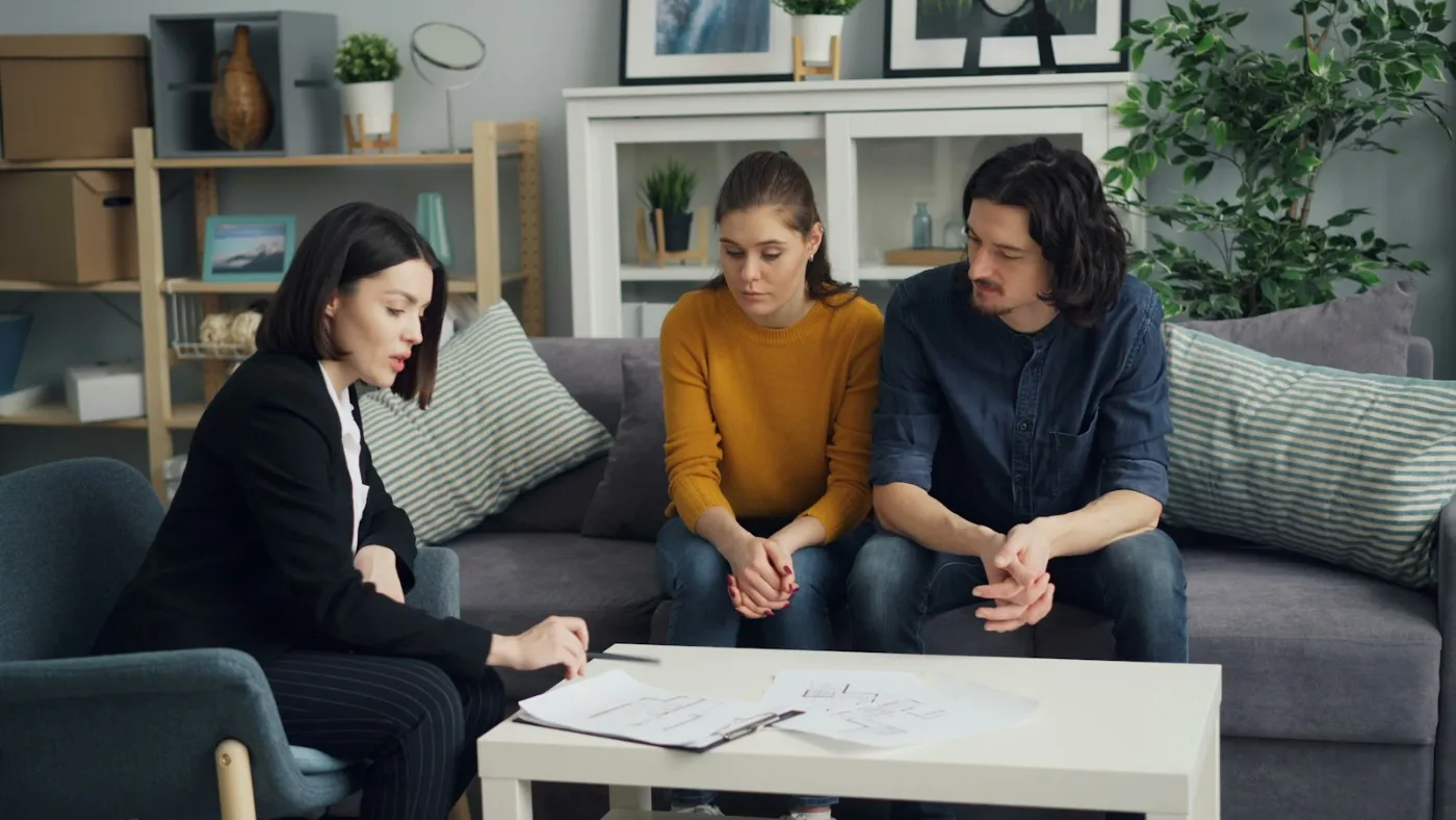 Corretora conversa com um casal na sala de estar, com documentos sobre a mesa